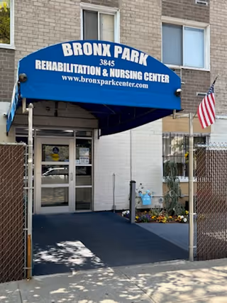 Entrance of Bronx Park Rehabilitation & Nursing Center with a blue awning showing the facility name and website, a gated walkway, and an American flag.