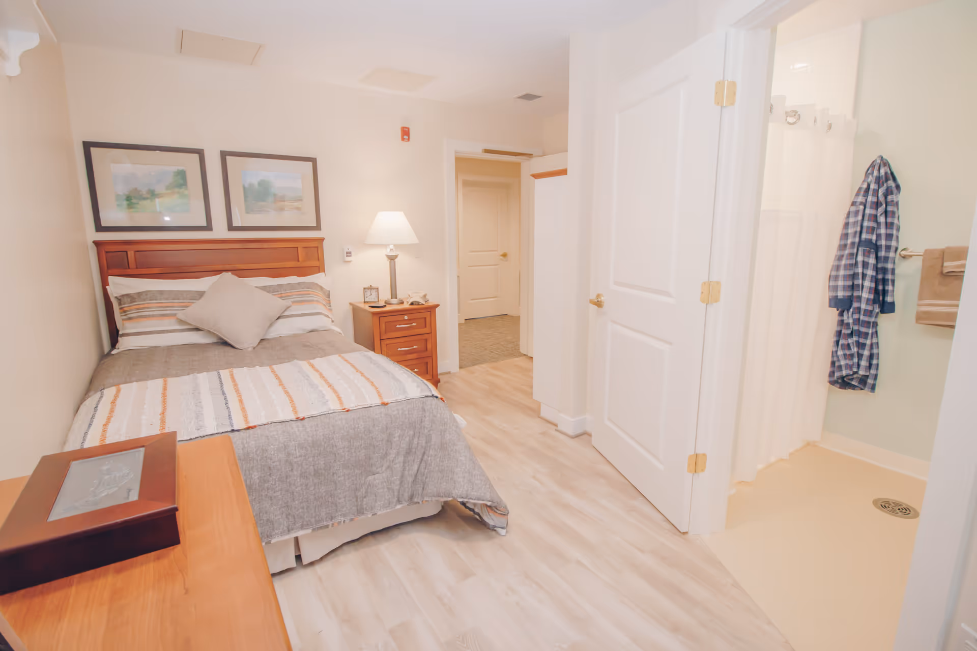 A cozy bedroom in a senior living facility featuring a neatly made bed with striped bedding and a pillow, a wooden nightstand with a lamp and clock, two framed landscape paintings on the wall, light wood flooring, and an open door leading to a bathroom with a shower and a hanging plaid robe.