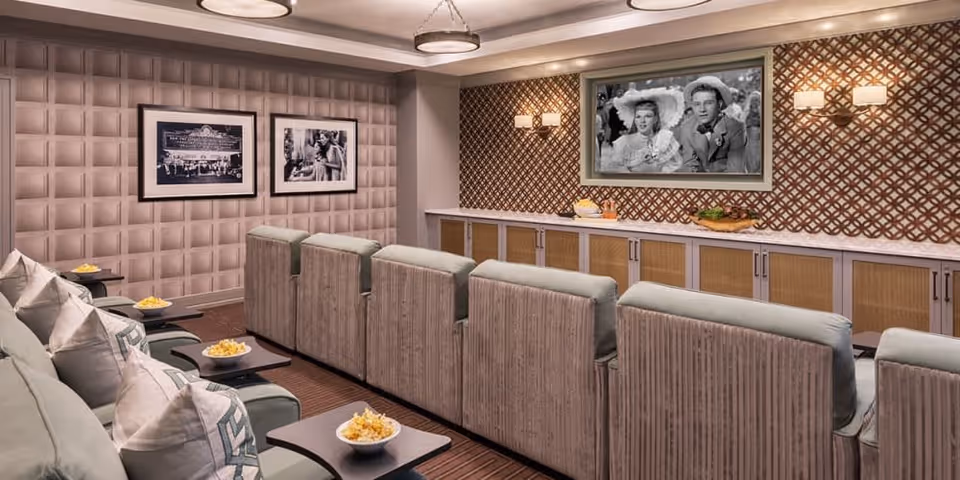 A cozy movie theater room with two rows of comfortable reclining chairs, each with a small table holding a bowl of popcorn. The walls are decorated with framed black and white photos and a patterned accent wall behind a large screen showing a black and white movie scene. Soft lighting fixtures are mounted on the walls, creating a warm ambiance.