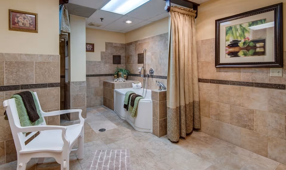 A spacious bathroom with beige and brown tiled walls and floor. There is a white bathtub with a handrail and a showerhead, a beige shower curtain, and a white plastic chair with green and brown towels draped over it. The room is decorated with framed pictures and a small plant on the bathtub ledge.
