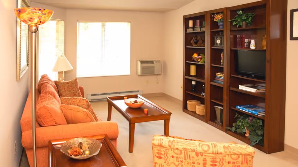 A cozy living room with an orange sofa and patterned armchair, a wooden coffee table with a bowl of decorative items, a tall floor lamp, a table lamp, and a large wooden bookshelf filled with books, plants, and decorative objects. The room has beige walls and carpet, and a window with blinds letting in natural light.