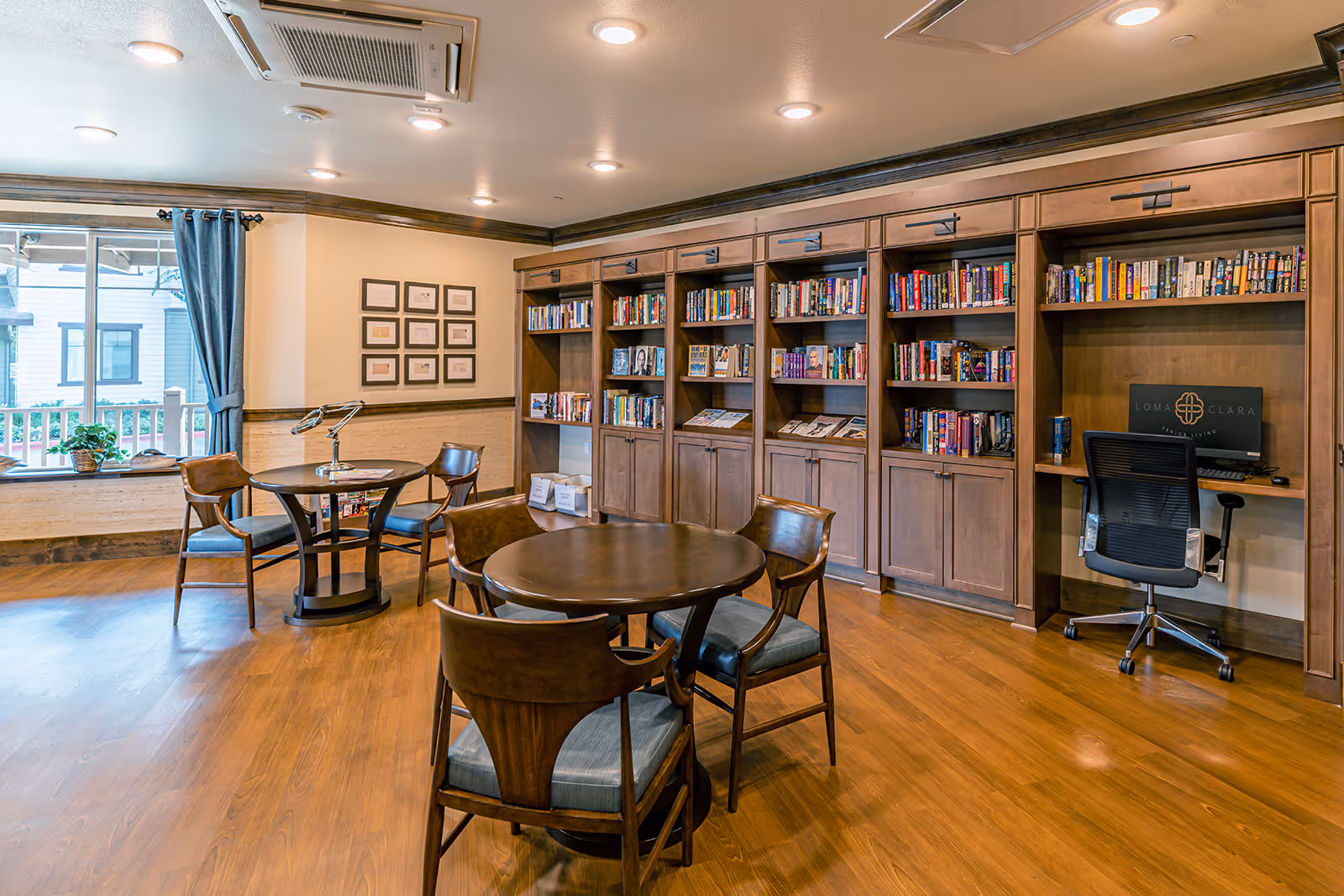 Well-lit senior living library/reading room with round tables and chairs in front of built-in wooden bookshelves and a small computer desk.