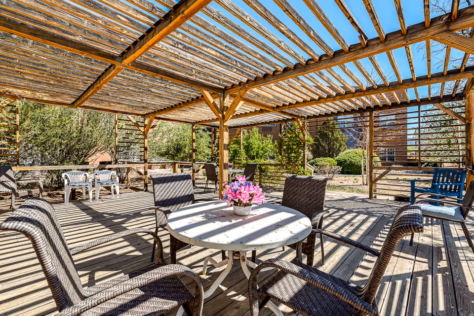 Shaded outdoor wooden pergola patio with wicker chairs around a round table topped with a pink flower arrangement.