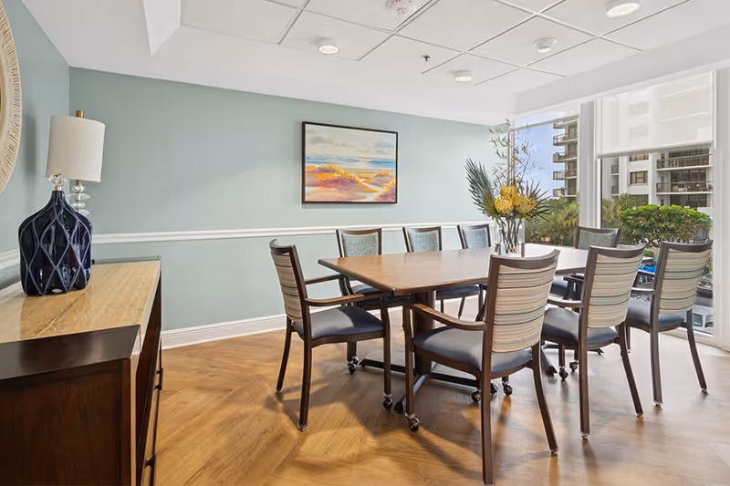 A bright dining room with a wooden table surrounded by eight chairs with blue cushions. A vase with yellow flowers and green leaves is placed on the table. The room has light blue walls with white trim, a wooden sideboard with a blue lamp, a framed landscape painting on the wall, and large windows showing an outdoor view of buildings and greenery.