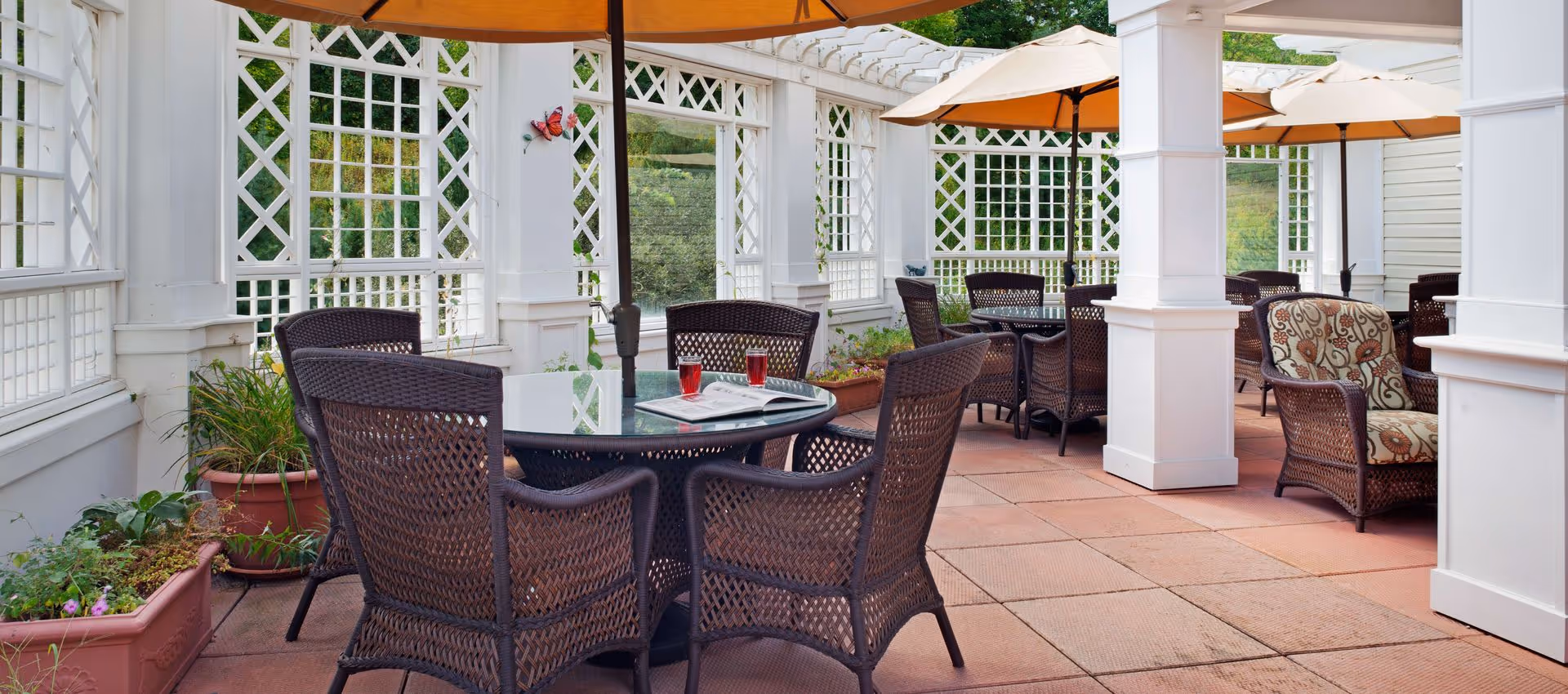 Outdoor patio area with multiple round glass tables surrounded by wicker chairs. Large beige umbrellas provide shade, and there are white lattice walls with greenery visible outside. Potted plants and a butterfly decoration are also present.