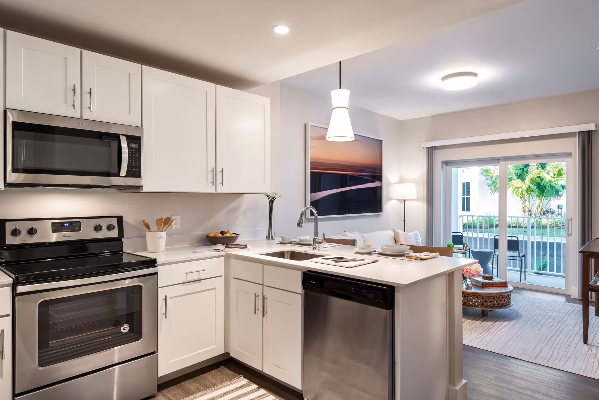 Modern kitchen with white cabinets, stainless steel appliances including a microwave, oven, and dishwasher. The kitchen has a white countertop with a sink and faucet, and is open to a living area with a sliding glass door leading to an outdoor patio with chairs and greenery. The living area has a floor lamp, a framed picture on the wall, and a small round coffee table with books and flowers.