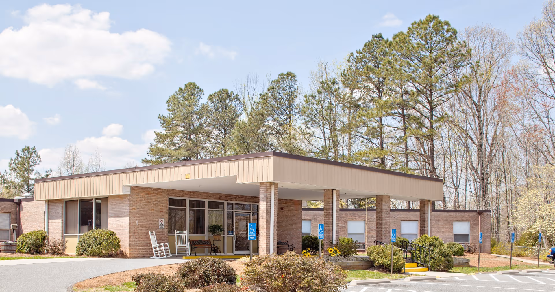 Exterior view of a single-story brick building with a covered entrance, surrounded by trees and bushes under a partly cloudy sky. There are rocking chairs and benches near the entrance and several handicap parking signs in the parking lot.