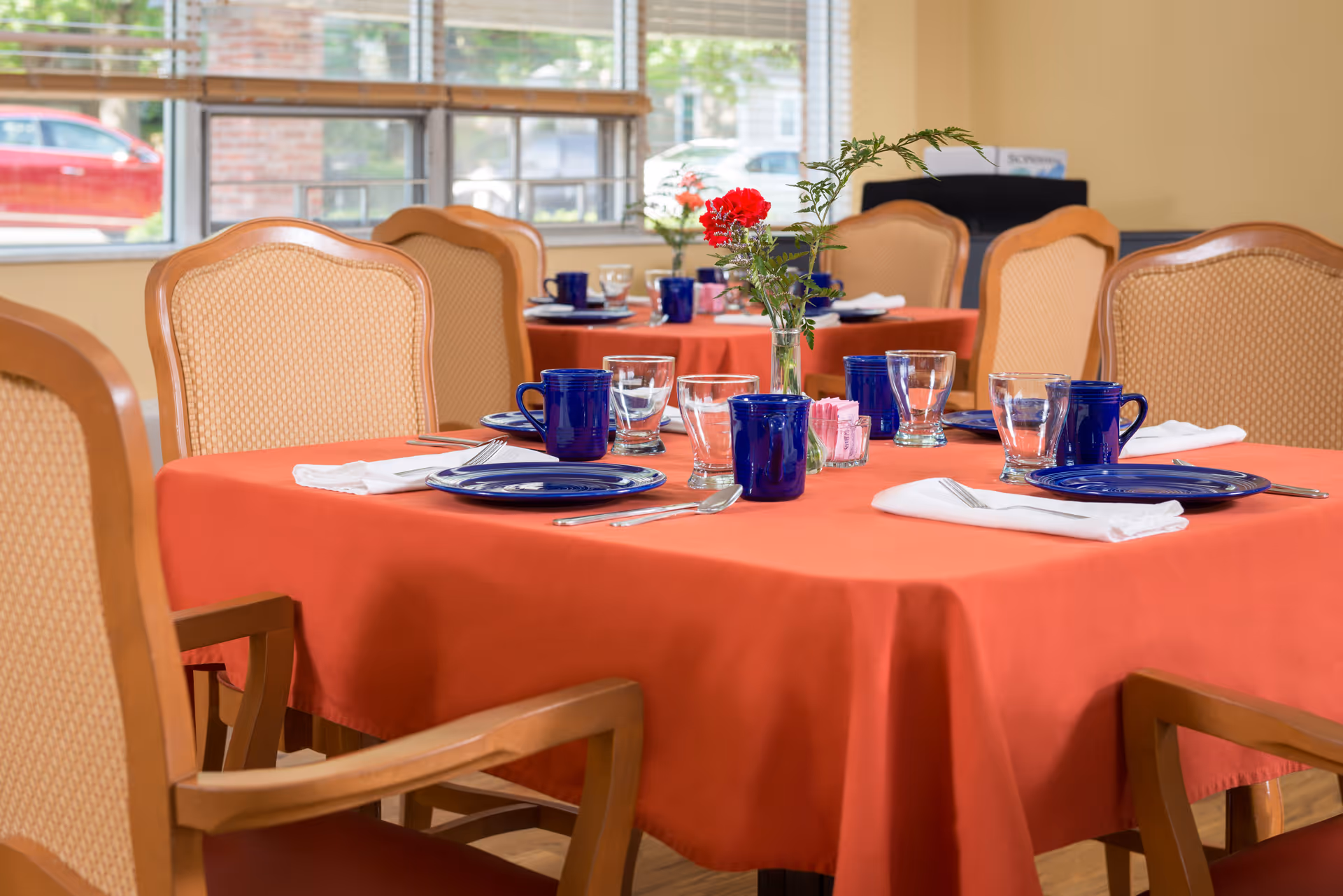 A dining room table set with blue plates, blue mugs, clear drinking glasses, silverware wrapped in white napkins, and a small vase with red and green flowers. The table is covered with an orange tablecloth and surrounded by wooden chairs with beige cushioned backs. Another similarly set table is visible in the background near a window.