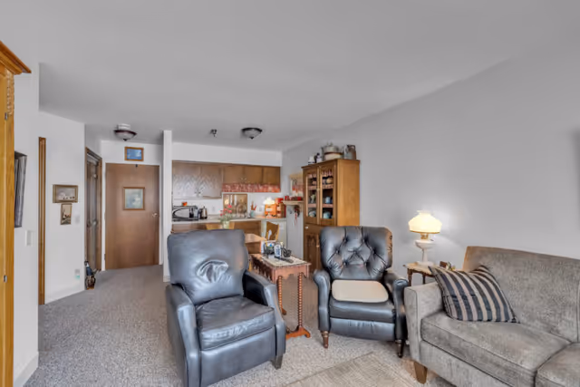 A cozy living room area in a nursing home with two black leather armchairs, a gray couch with a striped pillow, a wooden side table with a lamp, and a wooden cabinet filled with dishes. In the background, there is a small kitchen with wooden cabinets and a dining table with chairs.