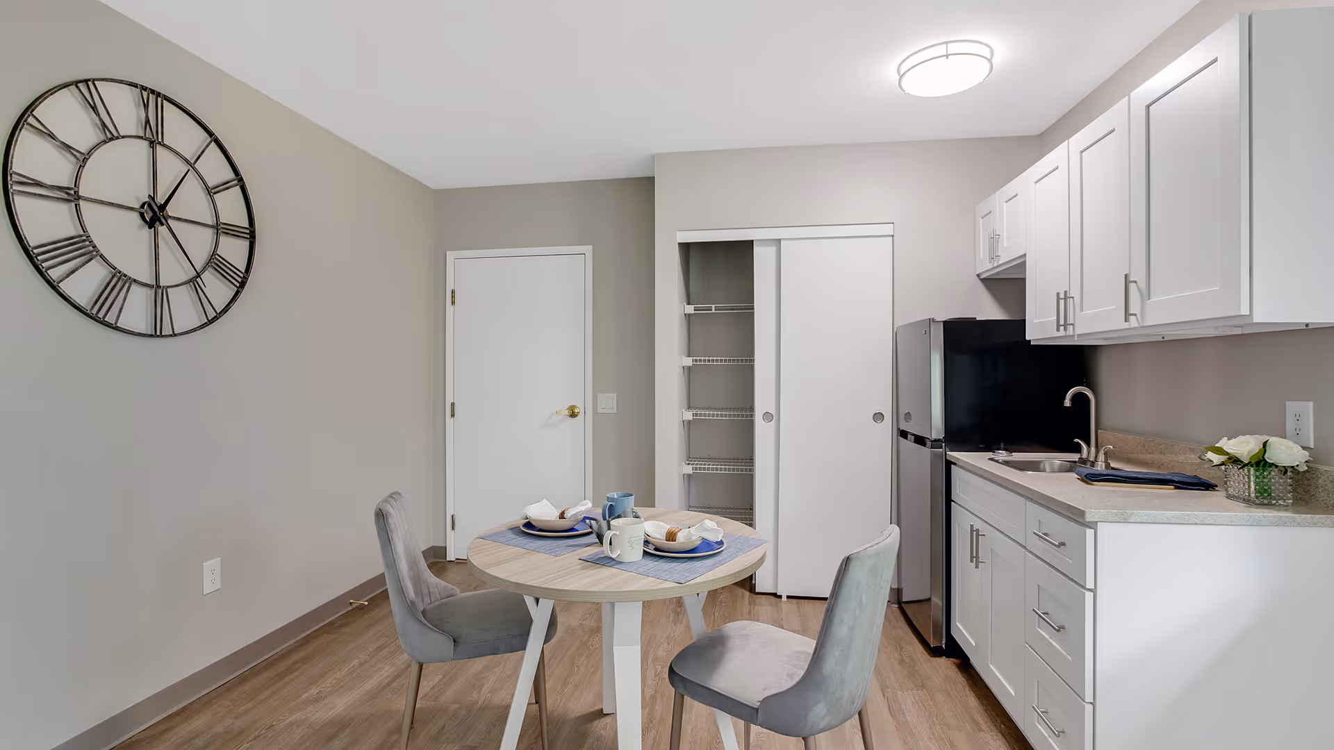 A small kitchen and dining area with a round table set for two, featuring plates, mugs, and napkins. The kitchen has white cabinets, a stainless steel refrigerator, a sink, and a countertop with a small flower arrangement. There is a large decorative wall clock on the beige wall, a closed white door, and a pantry with sliding doors.
