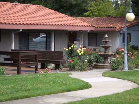Single-story senior living facility exterior with curved walkway, benches, fountain, and garden.