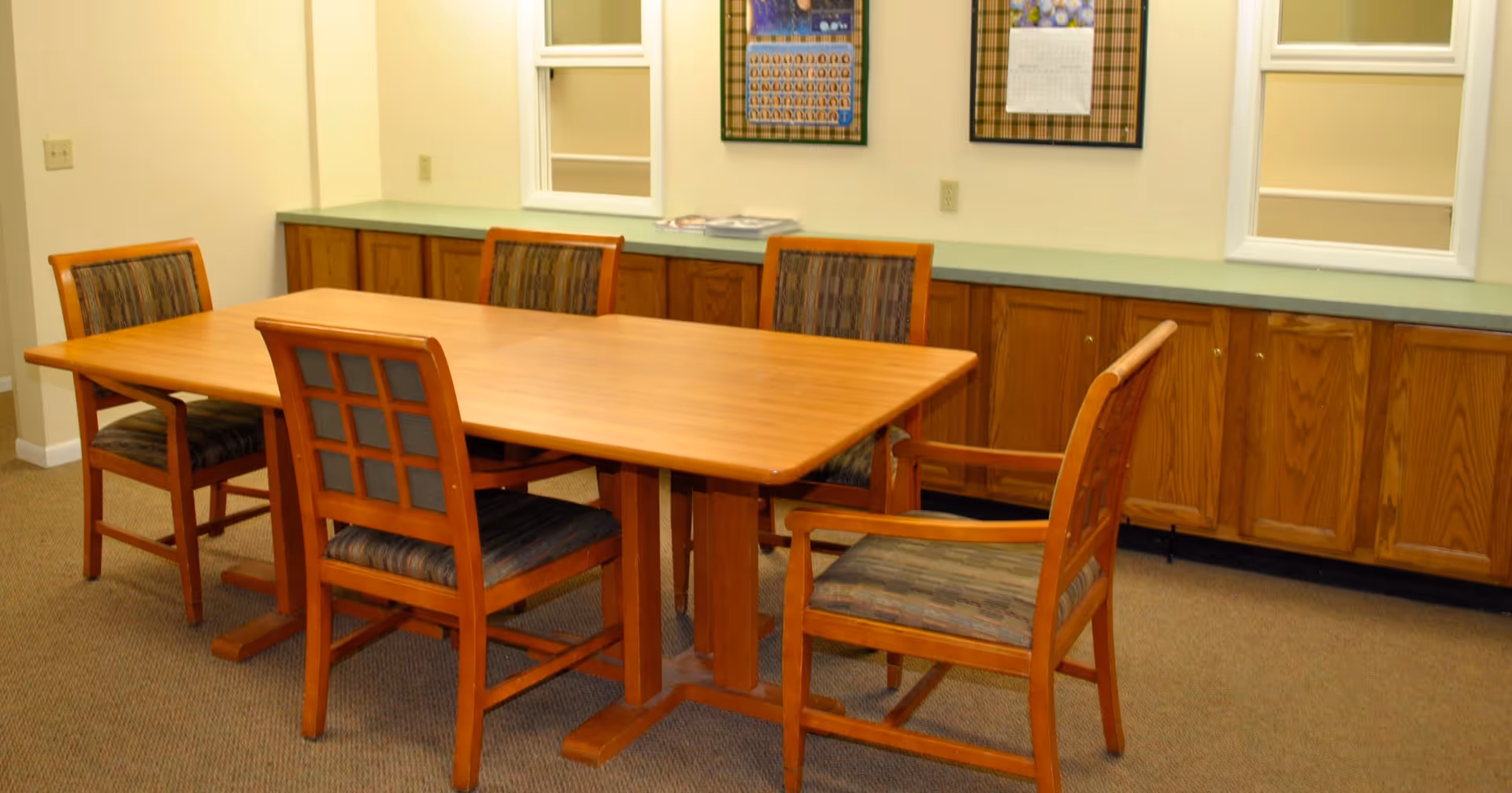 A room with a rectangular wooden table surrounded by five wooden chairs with cushioned seats and backs. The room has beige walls, a carpeted floor, and a long wooden cabinet with a green countertop along the back wall. Two framed calendars or posters are hanging on the wall above the cabinet.