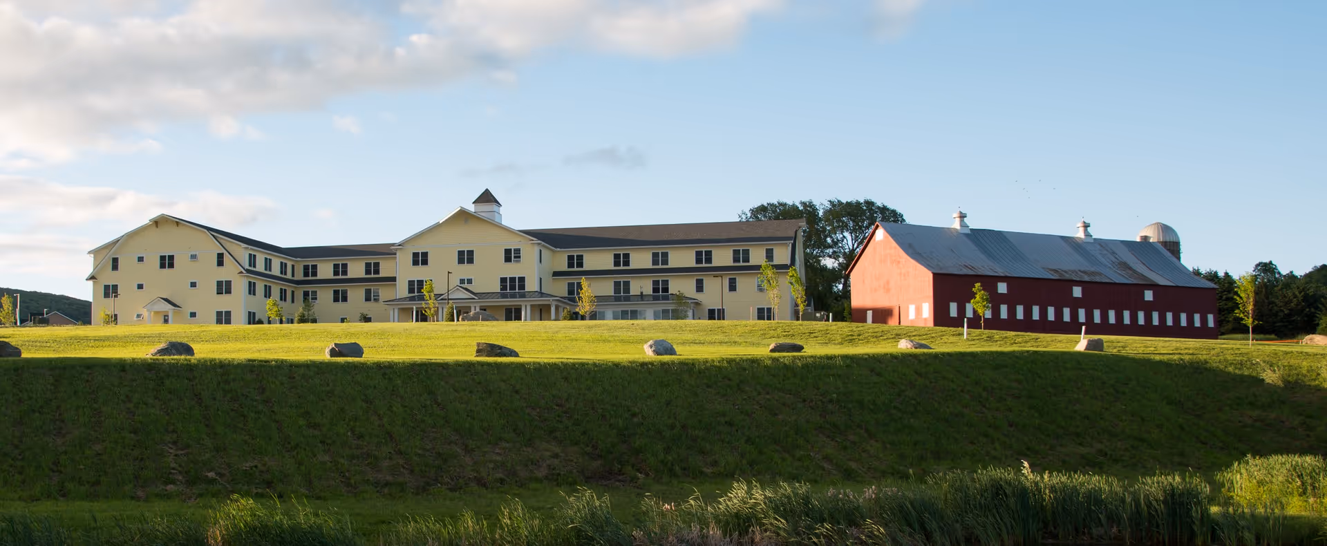 A large yellow senior living facility building with multiple windows and a pitched roof, situated on a grassy hill next to a red barn with a metal roof. The sky is partly cloudy with blue patches.