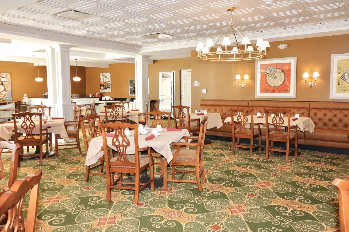 A spacious dining room with multiple wooden tables and chairs arranged neatly. The tables are covered with beige tablecloths and set with napkins and condiments. The room features a patterned green carpet, warm beige walls, framed artwork, and a large chandelier hanging from a decorative ceiling. There is a long cushioned bench along one wall and pendant lights in the background.