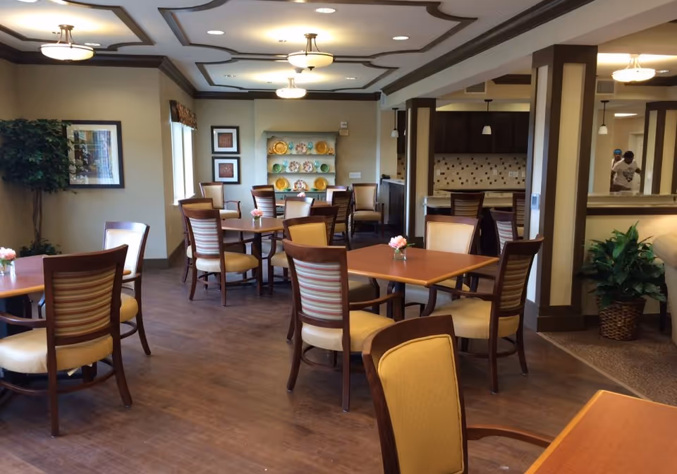 A dining room in a senior living facility with multiple wooden tables and cushioned chairs arranged neatly. The room features decorative ceiling beams, framed artwork on the walls, a shelf with colorful plates, and potted plants. The lighting is warm with ceiling fixtures, and there is a partial view of a kitchen area in the background.