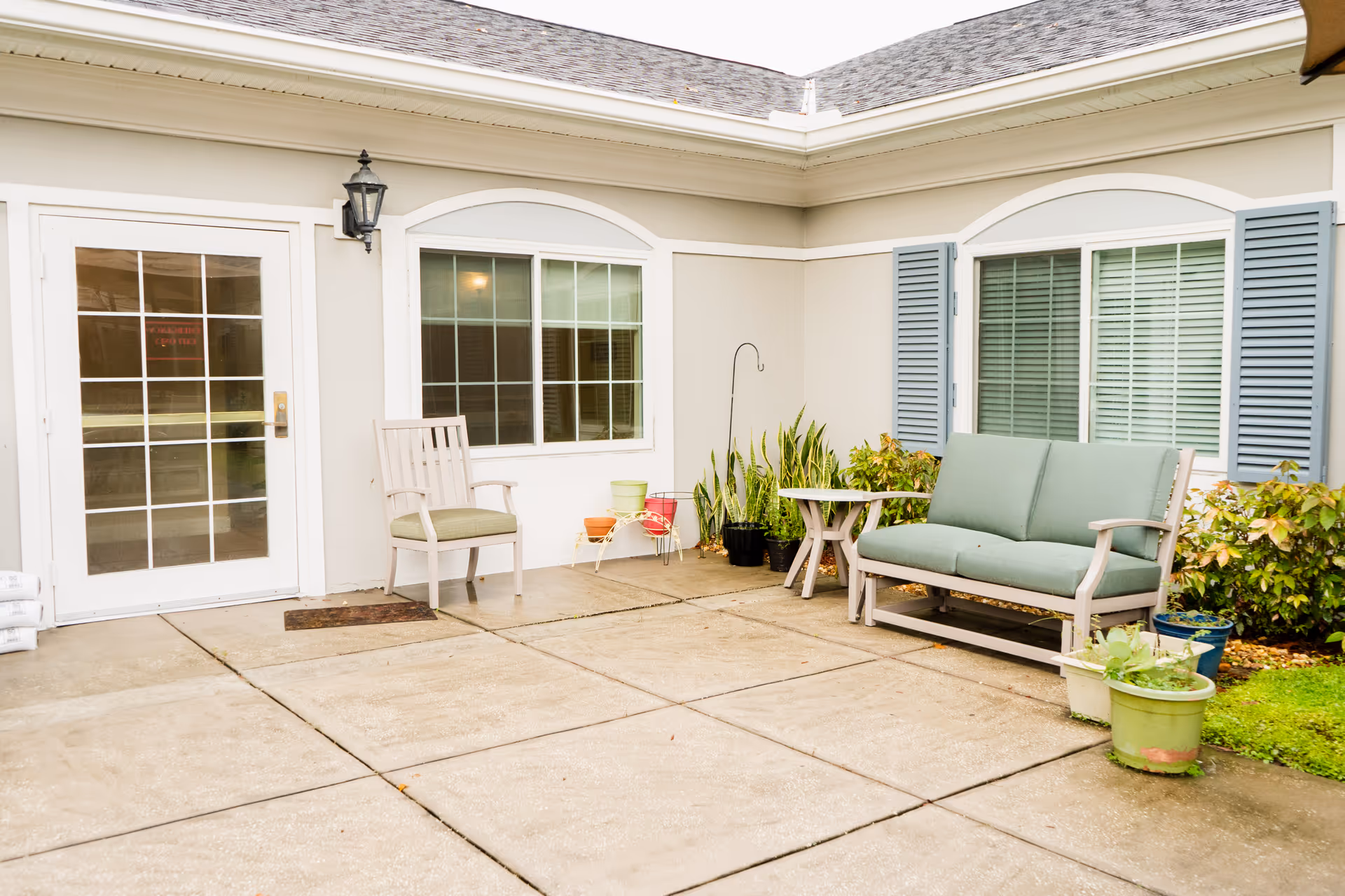 Outdoor patio area with a cushioned bench, a chair, a small table, several potted plants, and a glass door and window on the building wall.