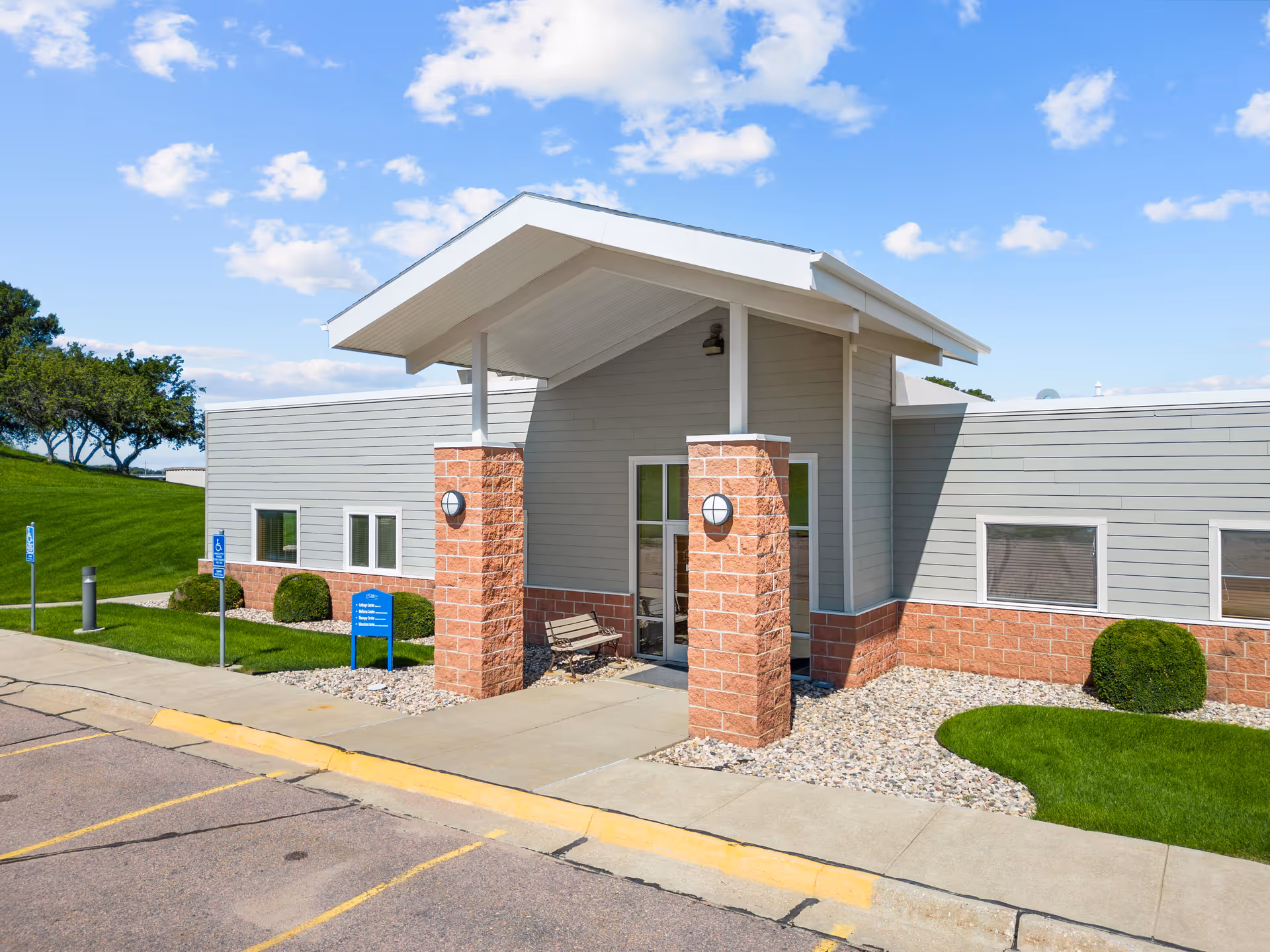 Exterior view of Good Samaritan Society - Le Mars building entrance with a covered porch supported by brick pillars, a bench, manicured bushes, green lawn, and a clear blue sky with scattered clouds.