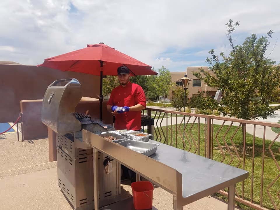 A man wearing a red shirt and black cap is grilling food on a large outdoor barbecue grill under a red umbrella. The setting is an outdoor patio area with a metal railing, green grass, trees, and beige buildings in the background under a partly cloudy sky.