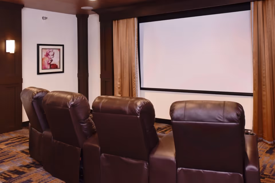 A small home theater room with four brown leather recliner chairs facing a large blank projection screen. The room has dark wood paneling, beige walls, a framed artwork on the left wall, and brown curtains on either side of the screen.