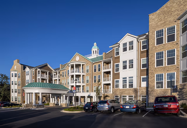 Exterior view of a multi-story senior living facility building with stone and beige siding, multiple windows, balconies, and a covered entrance with white columns. Several cars are parked in front under a clear blue sky.