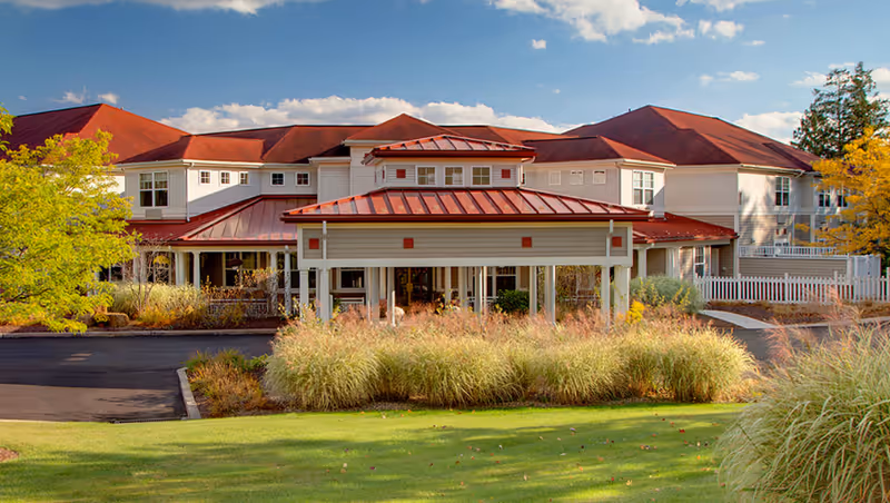 Exterior view of a senior living facility with a red roof, white walls, and a covered entrance. The building is surrounded by landscaped greenery, including grass, bushes, and trees under a partly cloudy blue sky.