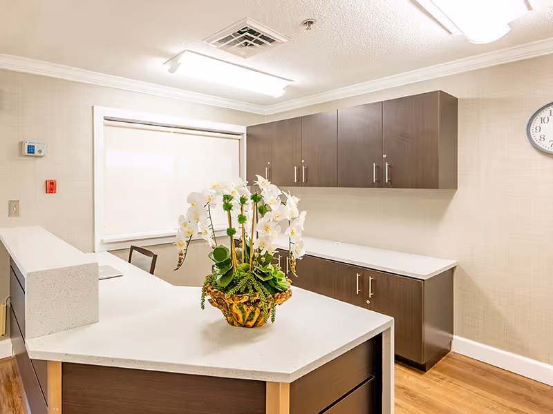 Interior view of a reception or front desk area with a white countertop and dark wood cabinets. A decorative plant with white flowers is placed on the counter. The room has beige walls, a clock on the wall, and fluorescent ceiling lights.