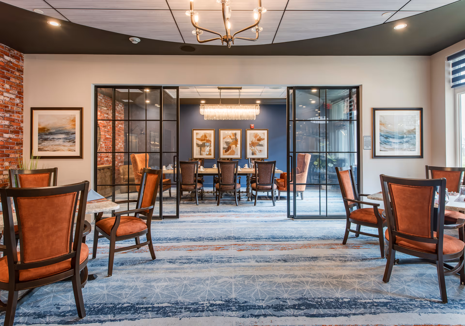 A spacious dining room with orange-upholstered chairs and tables facing a glass-partitioned private dining area and decorative artwork.