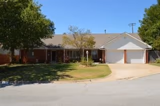 Single-story residential building with a brick and siding exterior, a two-car garage, and a driveway. The front yard has a lawn with some trees and shrubs, and the sky is clear and blue.