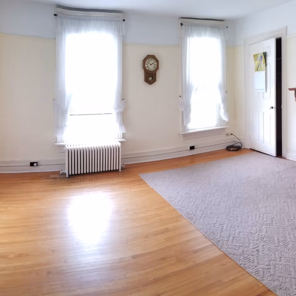 Empty sunlit living room with hardwood floors, two tall curtained windows with a radiator, a wall clock, a door, and a partially carpeted area.