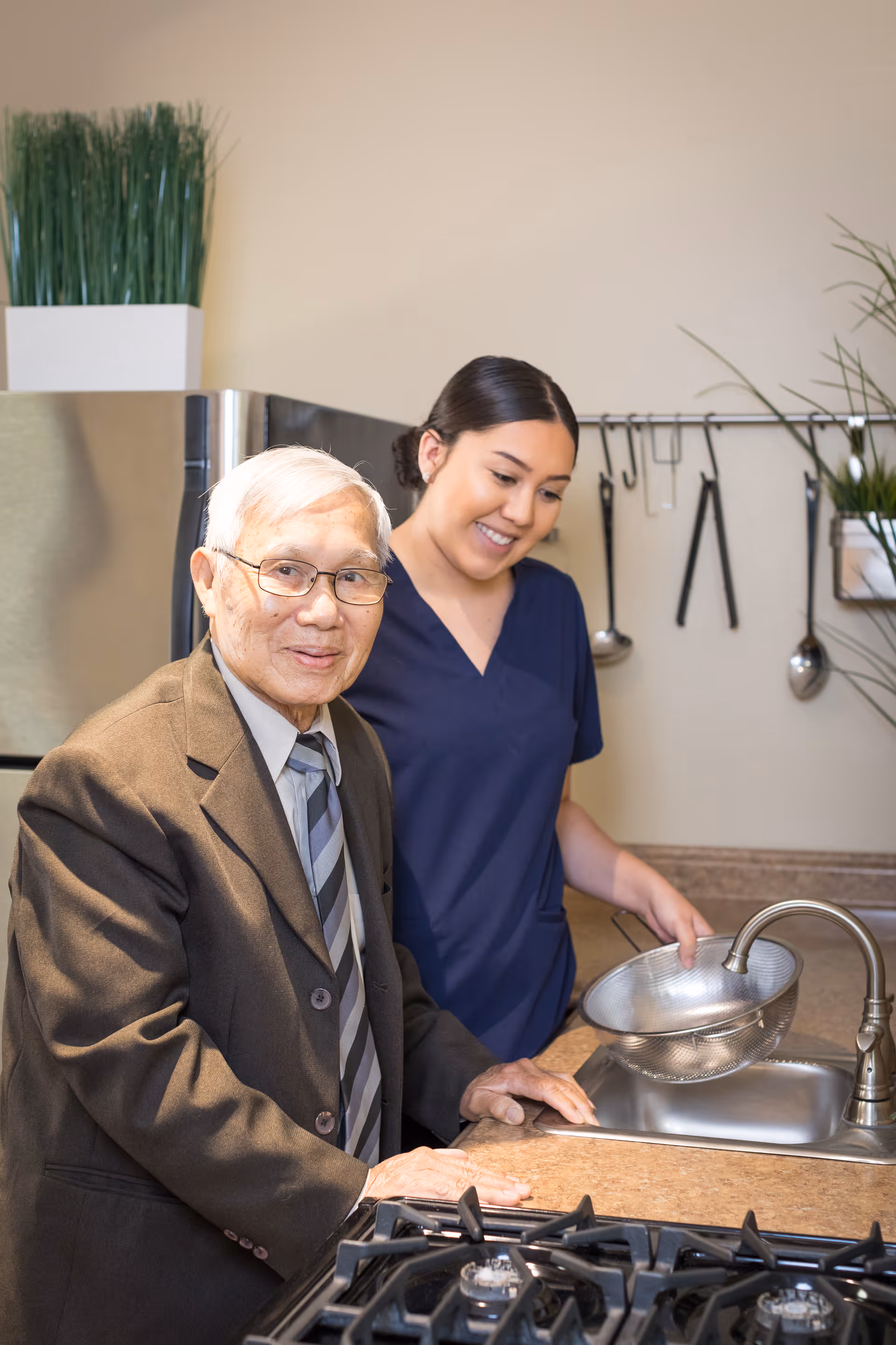 An elderly man wearing a suit and tie stands next to a younger woman in navy scrubs in a kitchen. The woman is holding a metal colander over the sink, and kitchen utensils hang on the wall behind them.