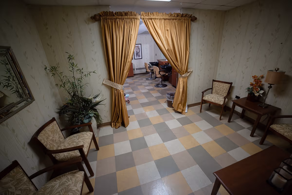 A waiting area with four patterned chairs, a small wooden table with a lamp and flower arrangement, and a potted plant. The floor has a checkered pattern with beige, gray, and light brown tiles. Gold curtains are tied back, revealing a room with salon chairs and mirrors.