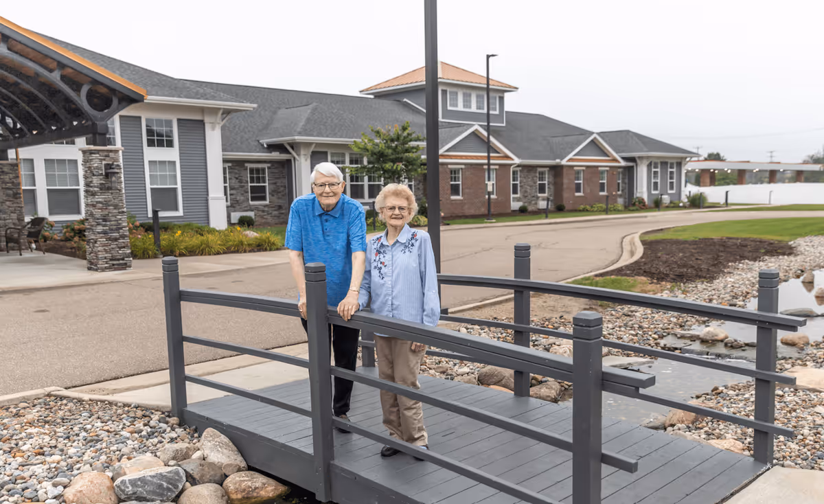 An elderly man and woman standing together on a small gray wooden bridge over a rock-lined stream in front of a senior living facility building with gray and brick exterior walls.