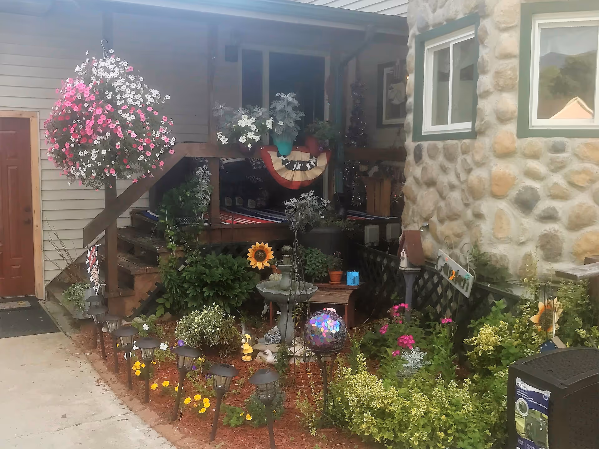 Outdoor garden area with various plants and flowers, including a hanging basket of pink and white flowers, a small birdbath, garden decorations, and a stone wall with two windows. There is a wooden staircase leading to a porch with more plants and decorations.