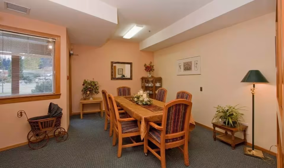 A dining room with a rectangular wooden table covered with a brown tablecloth and six wooden chairs with striped upholstery. The room has beige walls, a window with blinds showing an outdoor view, a small wooden side table with a floral arrangement, a corner cabinet with decorative items, a floor lamp with a green shade, and a small table with a potted plant.