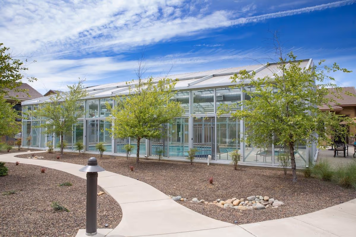 Outdoor view of a glass-enclosed swimming pool area at Eskaton Village Placerville, surrounded by a landscaped garden with small trees, shrubs, and a curved concrete walkway under a partly cloudy sky.