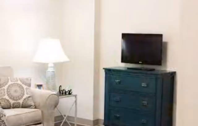 A cozy living room corner featuring a beige armchair with a patterned cushion, a white table lamp on a small side table, and a dark blue chest of drawers with a flat-screen TV mounted above it on a light-colored wall.