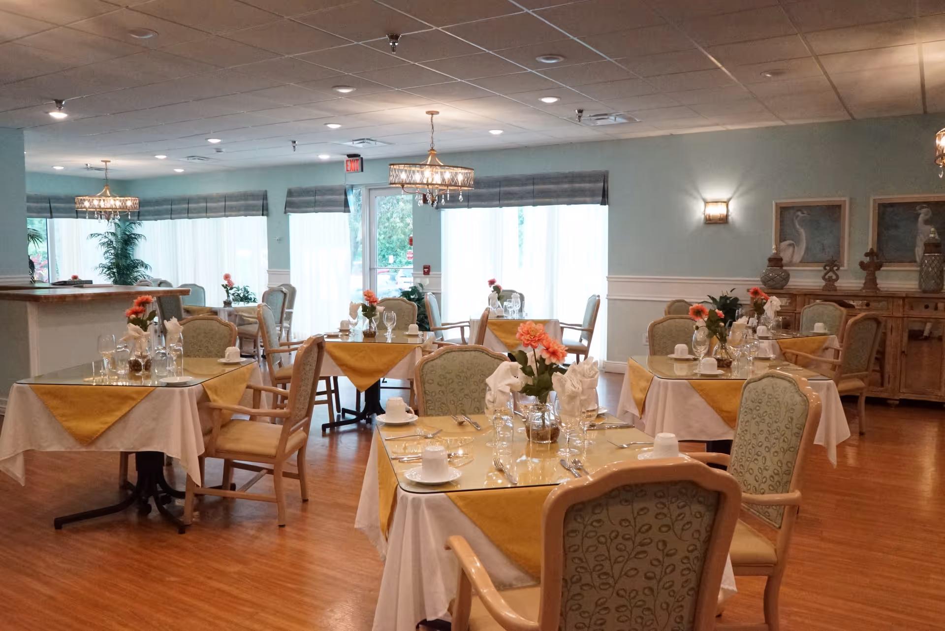 A bright and elegant dining room with several tables covered in white tablecloths and yellow table runners. Each table is set with glassware, cups, napkins, and floral centerpieces. The room has wooden flooring, soft blue walls, large windows with sheer curtains, and decorative wall art. Comfortable upholstered chairs surround the tables, and chandeliers hang from the ceiling providing warm lighting.