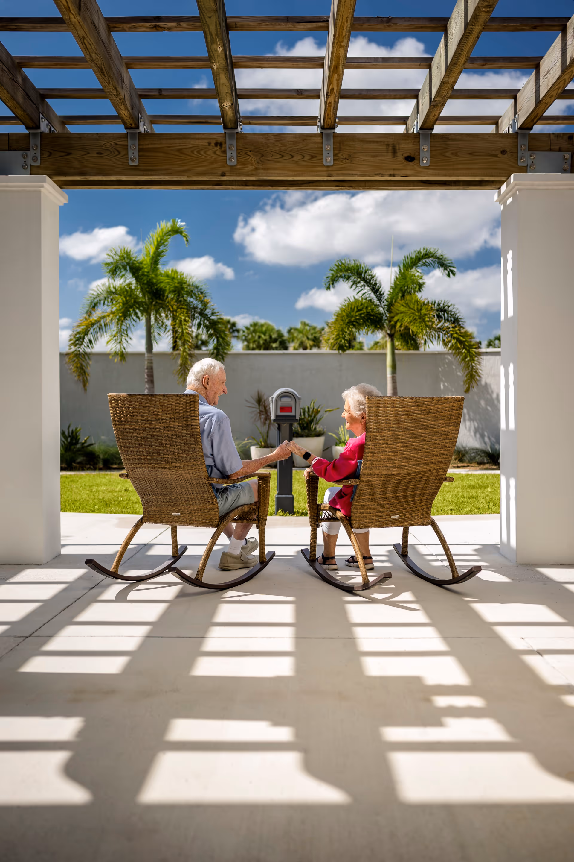 An elderly man and woman sitting in wicker rocking chairs under a wooden pergola, holding hands and facing each other with a backdrop of palm trees and a blue sky with clouds.
