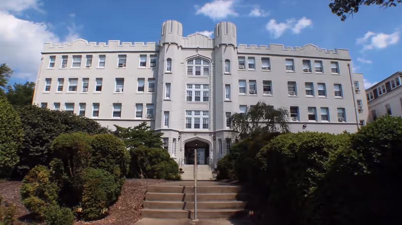 Front exterior view of a large, multi-story building with castle-like architectural features, including battlements and two cylindrical towers flanking the main entrance. The building is surrounded by well-maintained bushes and greenery, with a set of stairs leading up to the entrance under a partly cloudy blue sky.