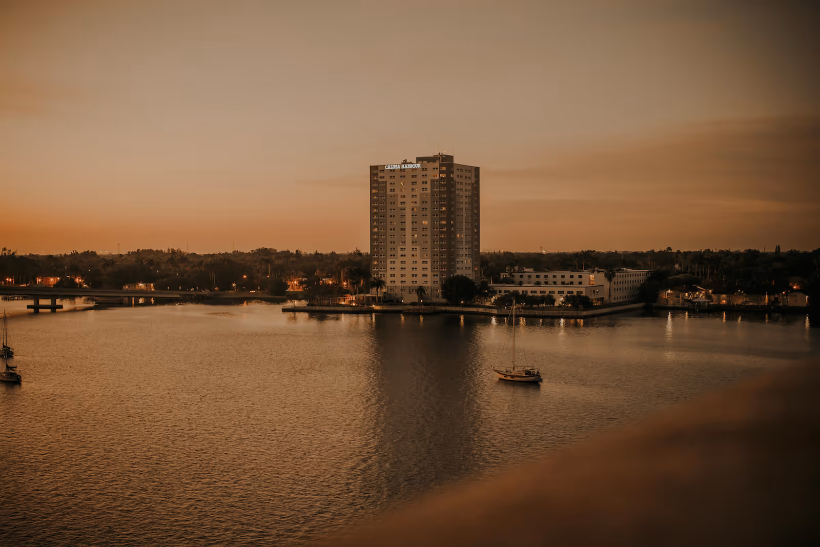 A tall building labeled Calusa Harbour stands near the edge of a body of water during sunset, with a sailboat floating nearby and a bridge visible in the background.
