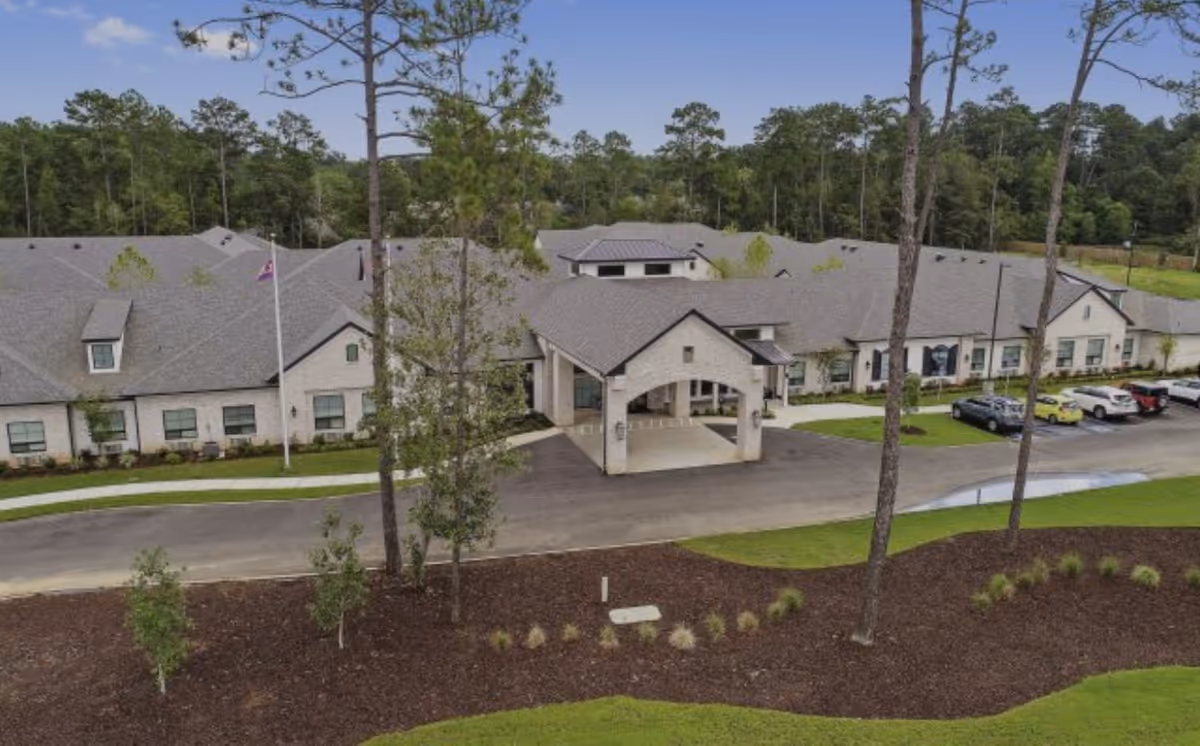 Aerial view of a single-story senior living facility building with a covered entrance, surrounded by trees and landscaped grounds. Several cars are parked in the parking lot adjacent to the building, and a flagpole is visible near the entrance.