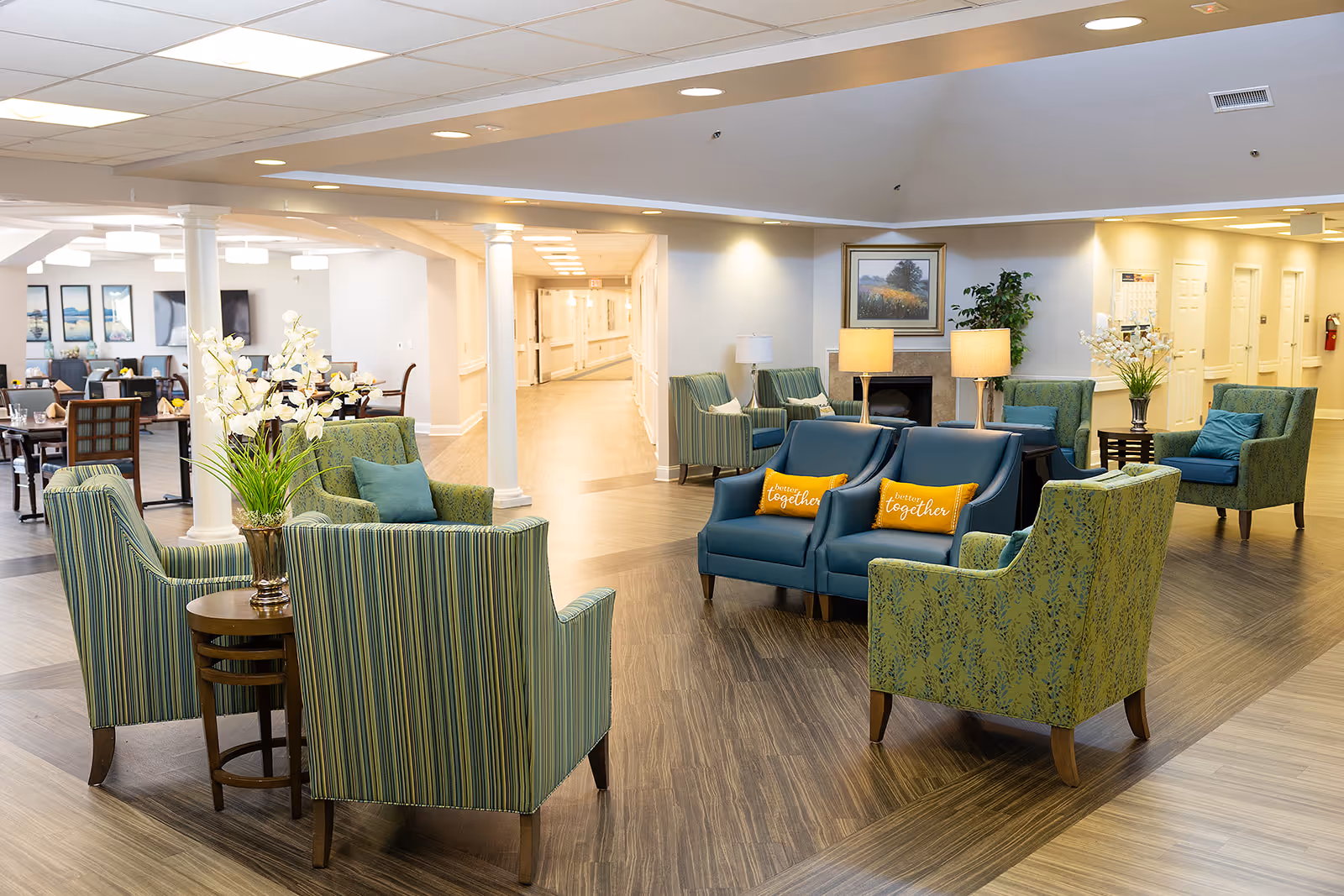 A bright and spacious senior living common area with multiple seating arrangements including green striped armchairs, teal armchairs with yellow pillows that say 'better together', and small tables with flower arrangements. The room features wood flooring, white columns, ceiling lights, a hallway leading further into the facility, and framed artwork on the walls.