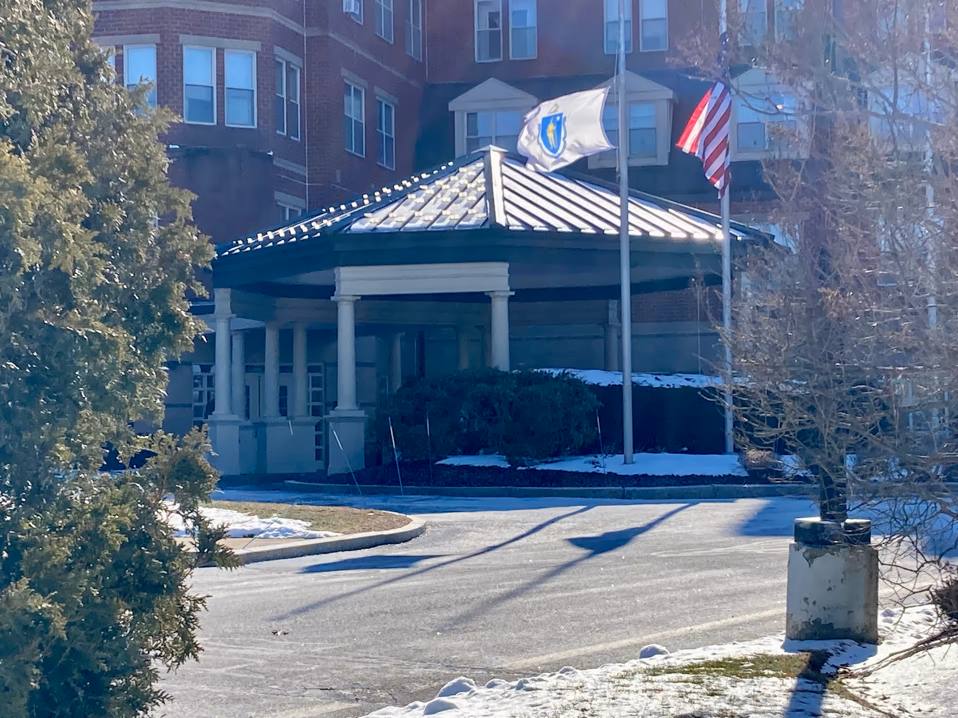 Front entrance of a brick senior living building with a covered porte-cochere, flagpoles, and light snow on the ground.