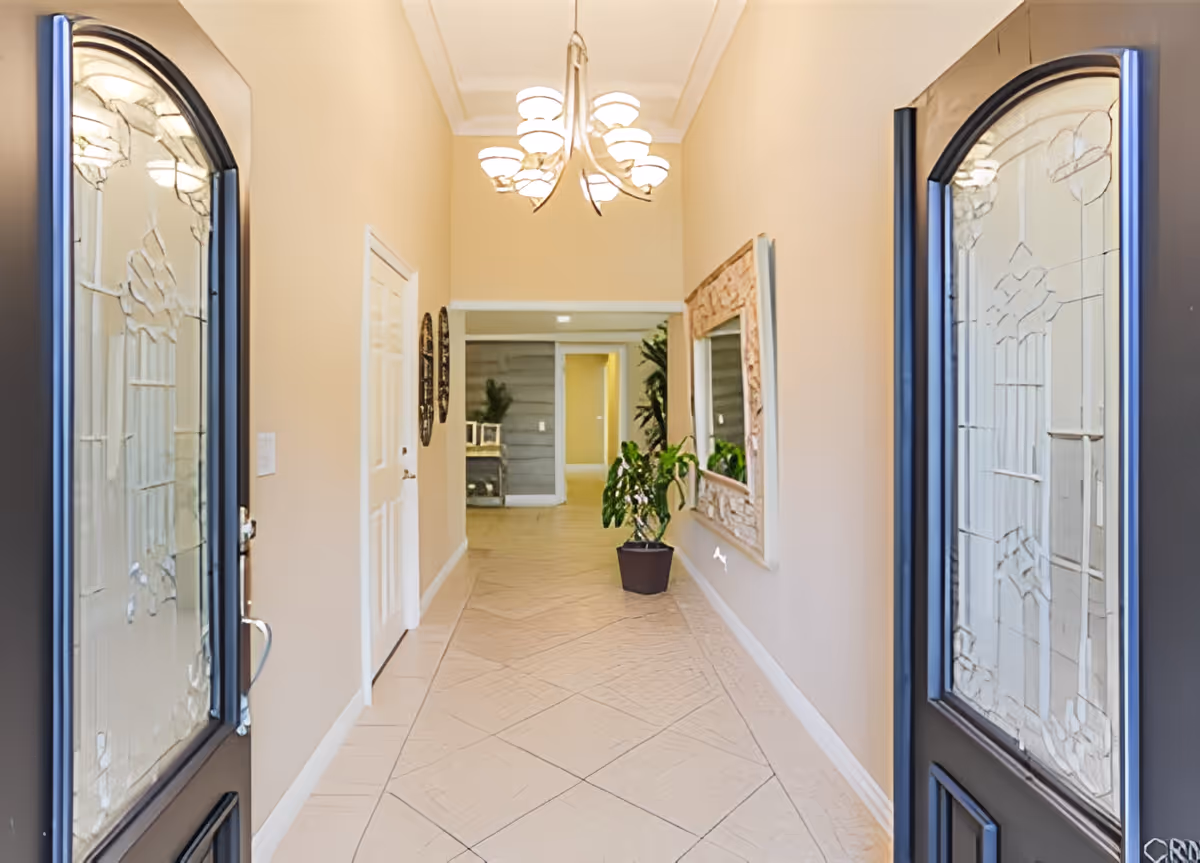 View through double front doors into a bright hallway with beige walls, tiled floor, a chandelier ceiling light, a large decorative mirror on the right wall, a potted plant beneath the mirror, and a small table with plants at the far end.