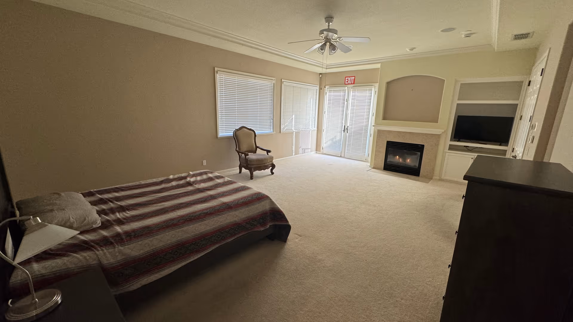 A spacious bedroom with a bed covered in a striped blanket, a wooden armchair near two windows with blinds, a ceiling fan, a fireplace, a built-in shelf with a TV, and a dresser. The room has beige walls and carpeted floor, with a glass door leading outside.