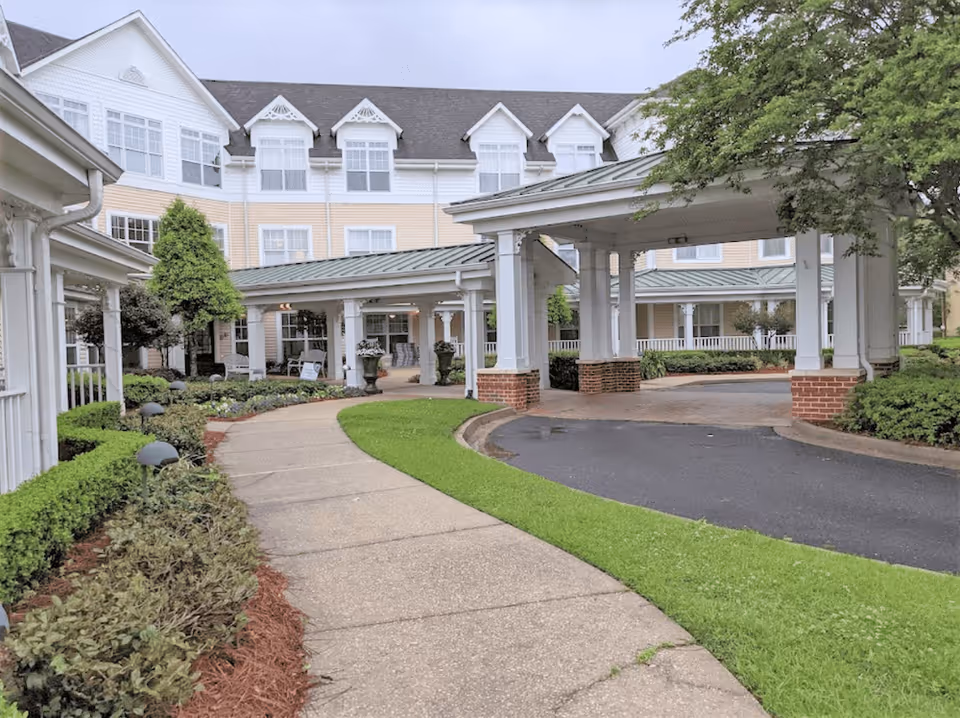 Exterior view of SummerHouse Vista Shores senior living facility showing a covered driveway entrance with white pillars and a green metal roof. The building is three stories tall with multiple windows and beige siding. There is a curved sidewalk leading to the entrance, surrounded by well-maintained landscaping including bushes, trees, and grass.