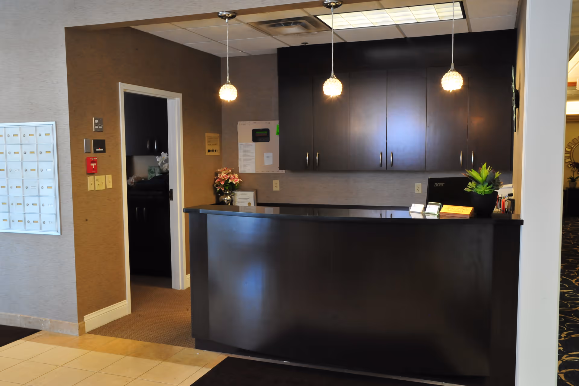 Reception desk area with dark wood cabinetry and counter, two hanging pendant lights, a computer, a small plant, and a flower arrangement. To the left is a wall with mailboxes and a doorway leading to another room.
