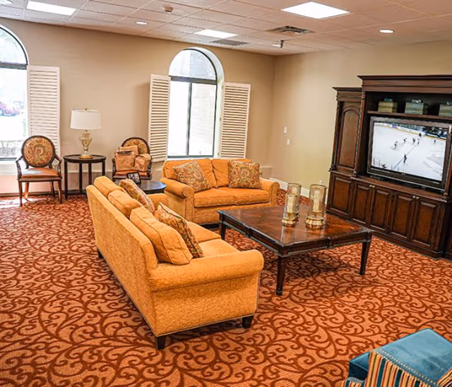 A cozy senior living facility lounge area with two orange sofas and patterned cushions arranged around a dark wooden coffee table. The room has a patterned carpet, two arched windows with white shutters, a wooden entertainment center with a TV showing a hockey game, and additional seating including chairs and a small side table with a lamp.
