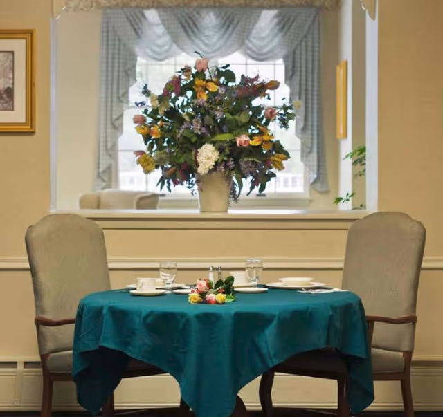 A small dining table covered with a teal tablecloth set for two with plates, cups, glasses, and a small floral centerpiece. Two upholstered chairs are positioned on either side of the table. Behind the table is a large window with sheer curtains and a large floral arrangement on the windowsill.