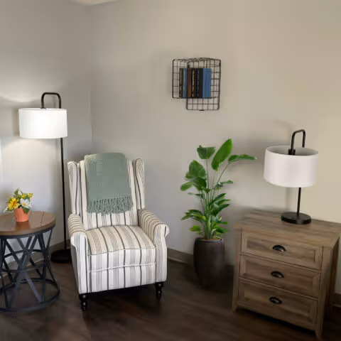A cozy corner of a living room featuring a striped armchair with a green throw blanket draped over it, a small round wooden side table with a potted yellow flower, a tall floor lamp, a wooden three-drawer chest with a table lamp on top, a green potted plant, and a small wall-mounted wire shelf holding books against a plain light-colored wall.